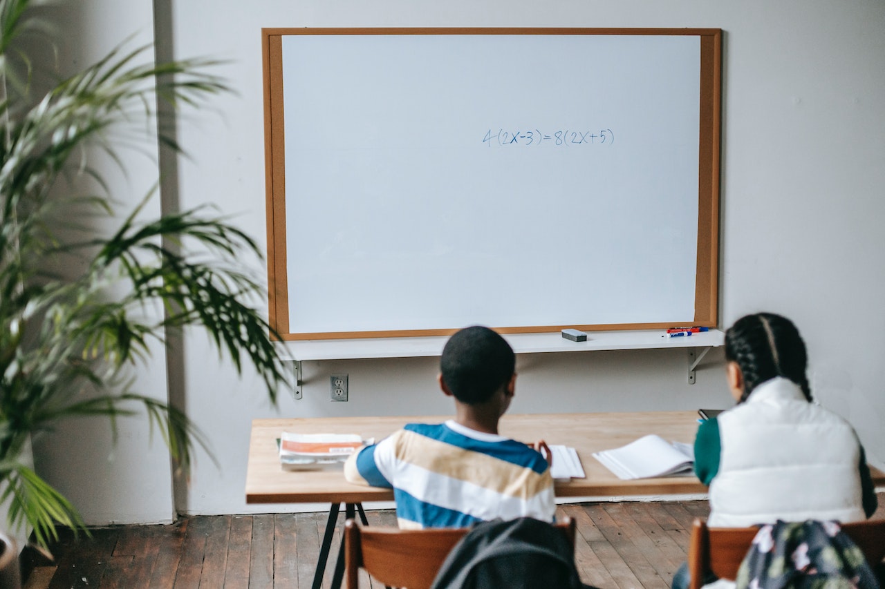 Two students sit at a wooden desk facing a whiteboard in a classroom. The whiteboard displays a mathematical equation, and there are various books and papers on the desk. A potted plant is partially visible in the left foreground.