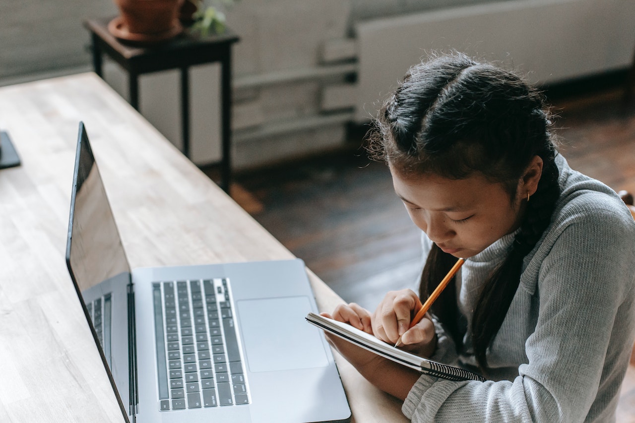 A young girl with braided hair sits at a wooden table, concentrating on writing in a notebook with a pencil. An open laptop is positioned in front of her. The background features a plant on a small table.
