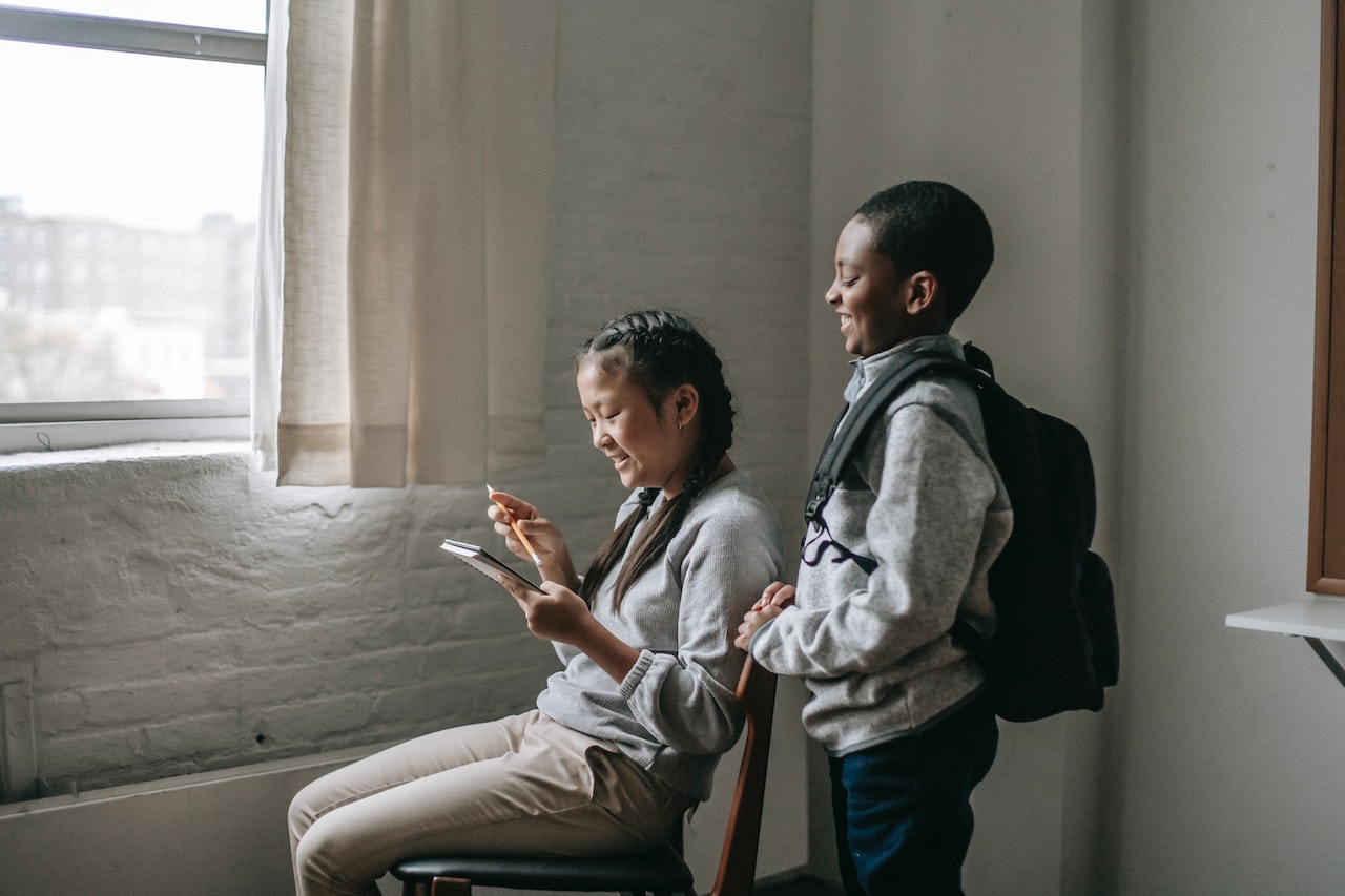 A girl with braided hair is sitting by a window, looking at her smartphone and smiling. Next to her stands a boy with a backpack, also smiling and looking at the phone. The scene appears to be indoors, in a room with light-colored walls.